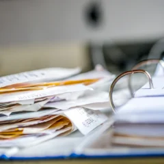 A close-up of a messy stack of papers and receipts next to an open ring binder on a desk, with a blurred background.