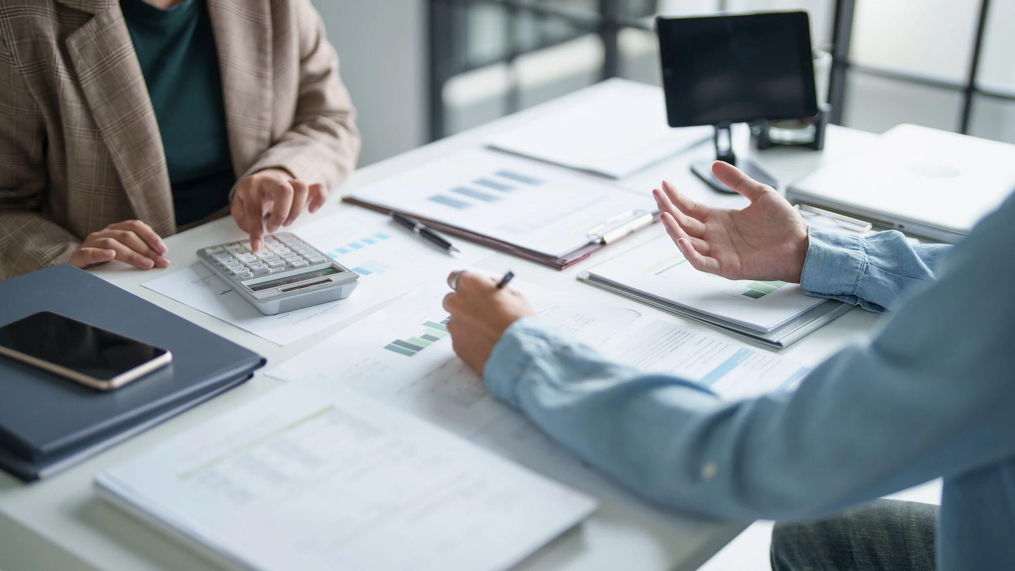 Two people sit at a desk with charts, graphs, a calculator, and documents, discussing financial data. One gestures with their hands while the other types on a calculator. Laptops and papers are spread across the workspace.