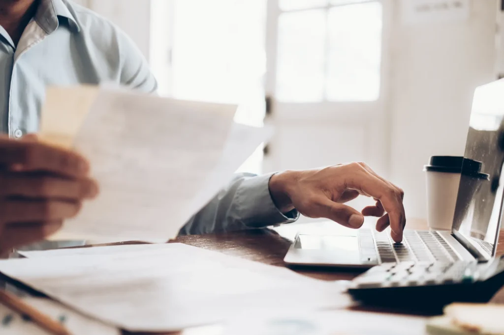 Person in a light blue shirt holds documents in one hand and types on a laptop with the other. Papers, a coffee cup, and a calculator are on the wooden desk. Sunlight streams through a window in the background.