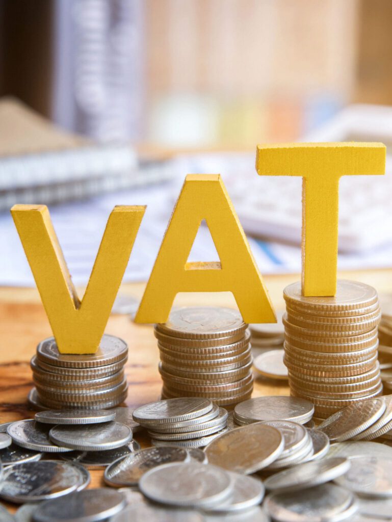 Yellow letters spelling “VAT” sit on stacks of coins, with more coins scattered around and blurred paperwork in the background.