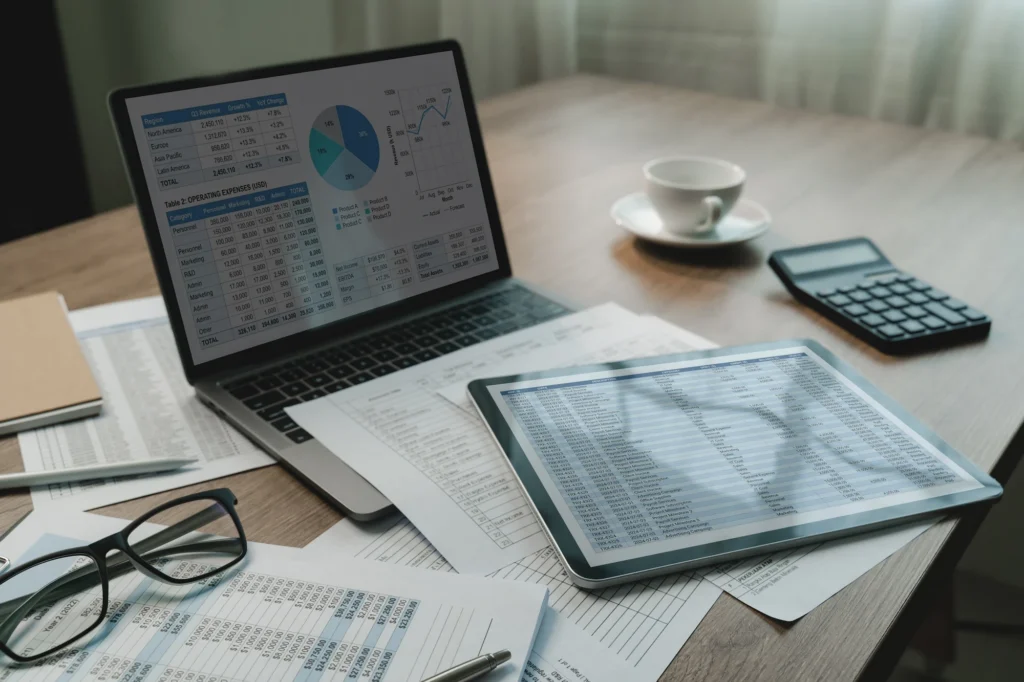 A laptop and tablet displaying spreadsheets sit on a desk with printed financial documents, eyeglasses, a calculator, and a coffee cup, suggesting a work setting focused on data analysis or accounting.