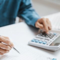 A person in a blue shirt writes on paperwork while using a calculator, with a computer keyboard in the background on a white desk.