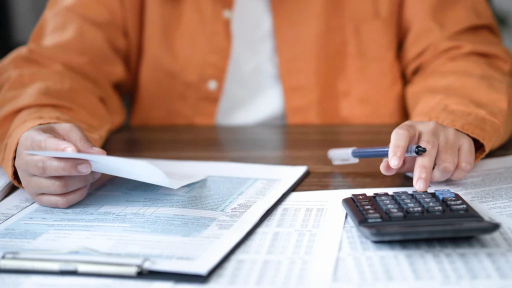 A person in an orange shirt reviews documents, holding a piece of paper in one hand and pressing buttons on a calculator with the other. Papers and charts are spread across the table.