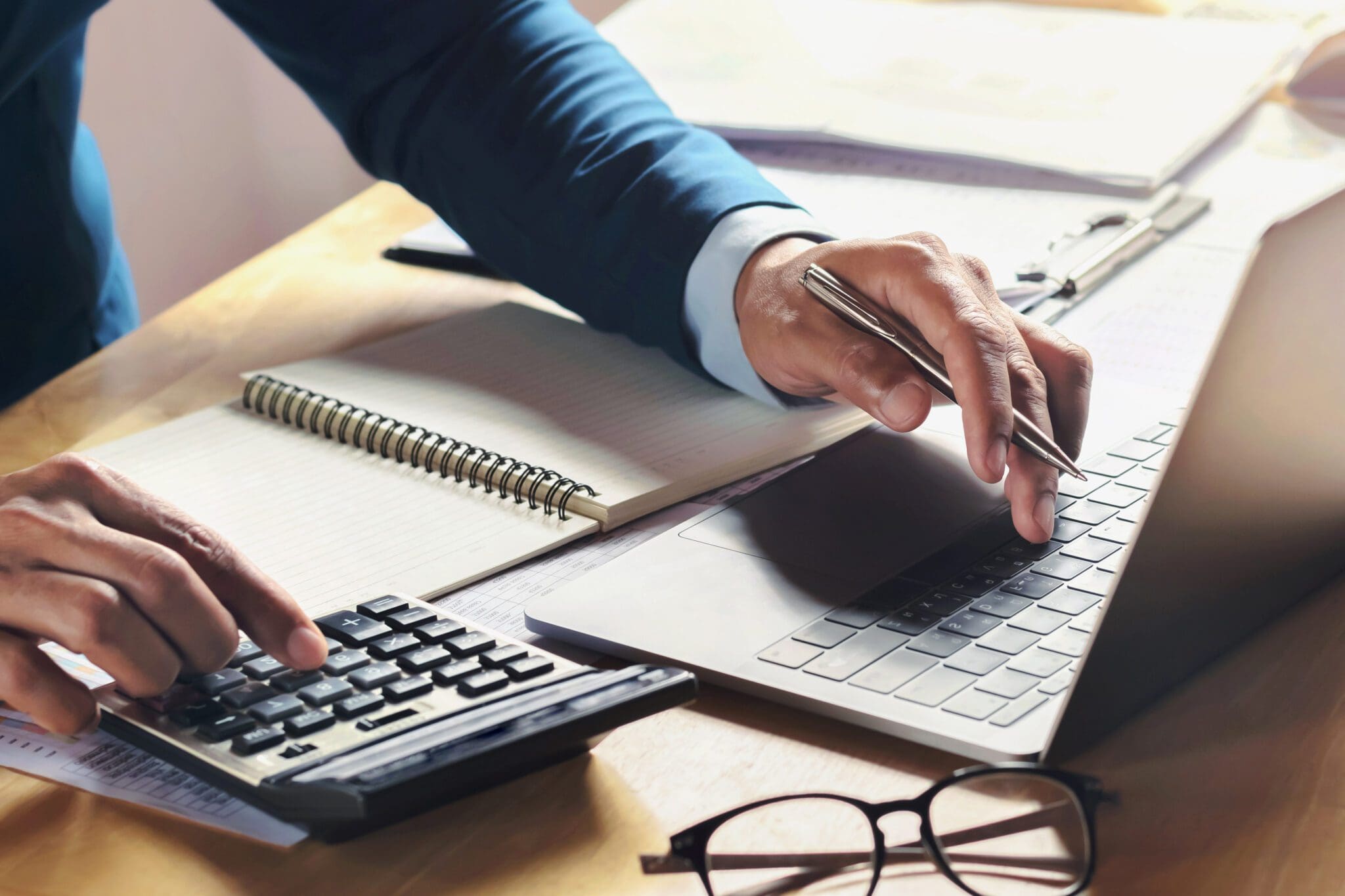 A person uses a calculator and types on a laptop at a desk with a notebook, papers, and eyeglasses nearby.