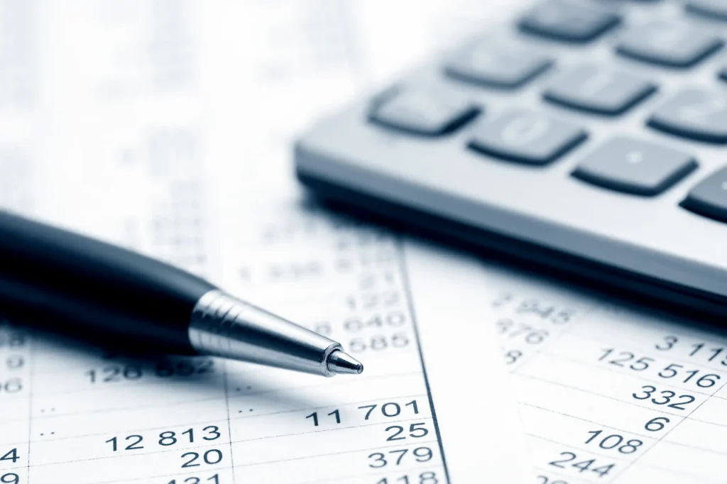 A close-up of a pen resting on a financial spreadsheet with numbers in rows and a calculator in the background, suggesting financial calculations or accounting work.