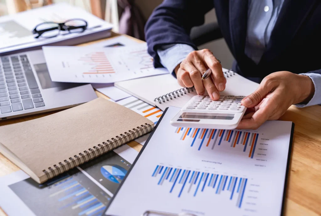 A person in business attire uses a calculator at a desk covered with financial charts, graphs, a laptop, notebooks, and papers.