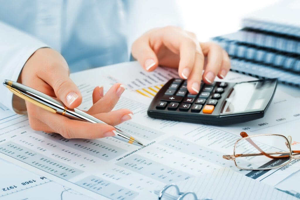 A person uses a calculator and holds a pen over financial documents, with eyeglasses and stacked papers nearby.