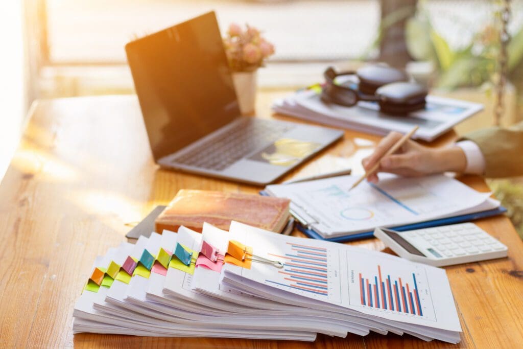 A person writes on papers with charts at a desk, surrounded by stacked reports, a laptop, a calculator, notebooks, and office supplies in a brightly lit workspace.