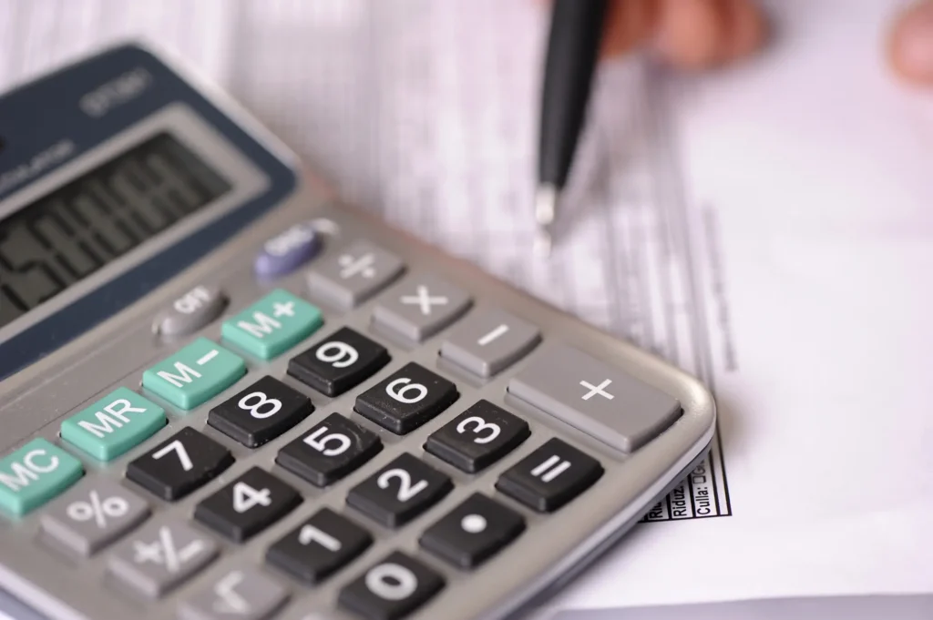 A close-up of a calculator on top of a document, with a hand holding a pen in the background, ready to write or make calculations.