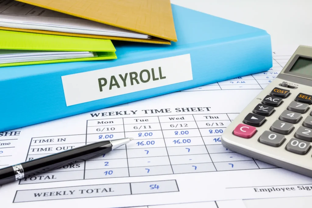 A calculator, pen, and folders labeled “PAYROLL” resting on a weekly time sheet, showing work hours and totals.