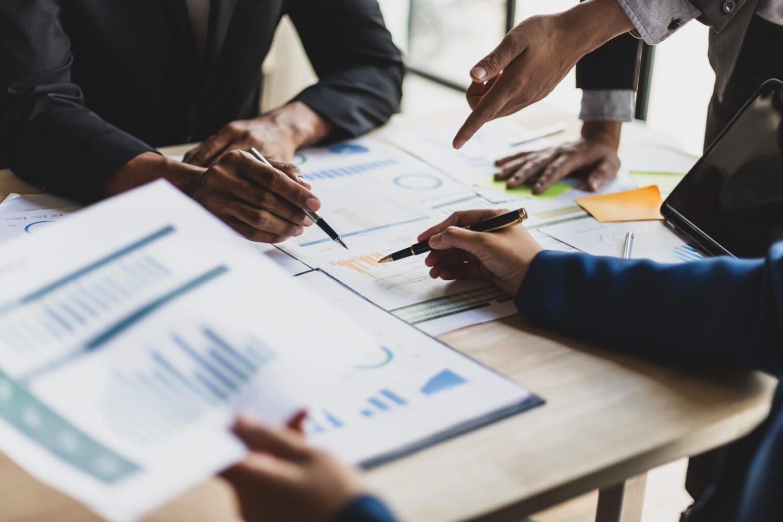 Close-up of several people’s hands reviewing and discussing financial charts and graphs on paper at a meeting table, with pens and documents visible.