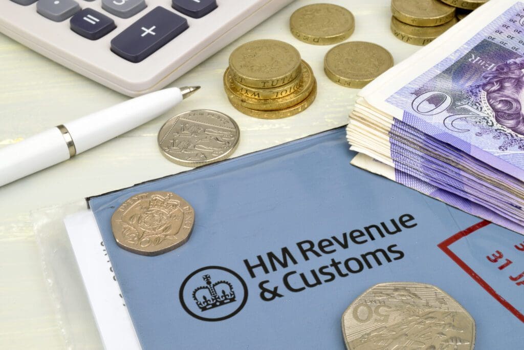 A folder labeled “HM Revenue & Customs” with British coins, banknotes, a white pen, and part of a calculator on a light desk.