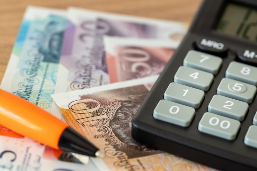 A close-up of British pound banknotes, a black and gray calculator, and an orange pen on a wooden surface.