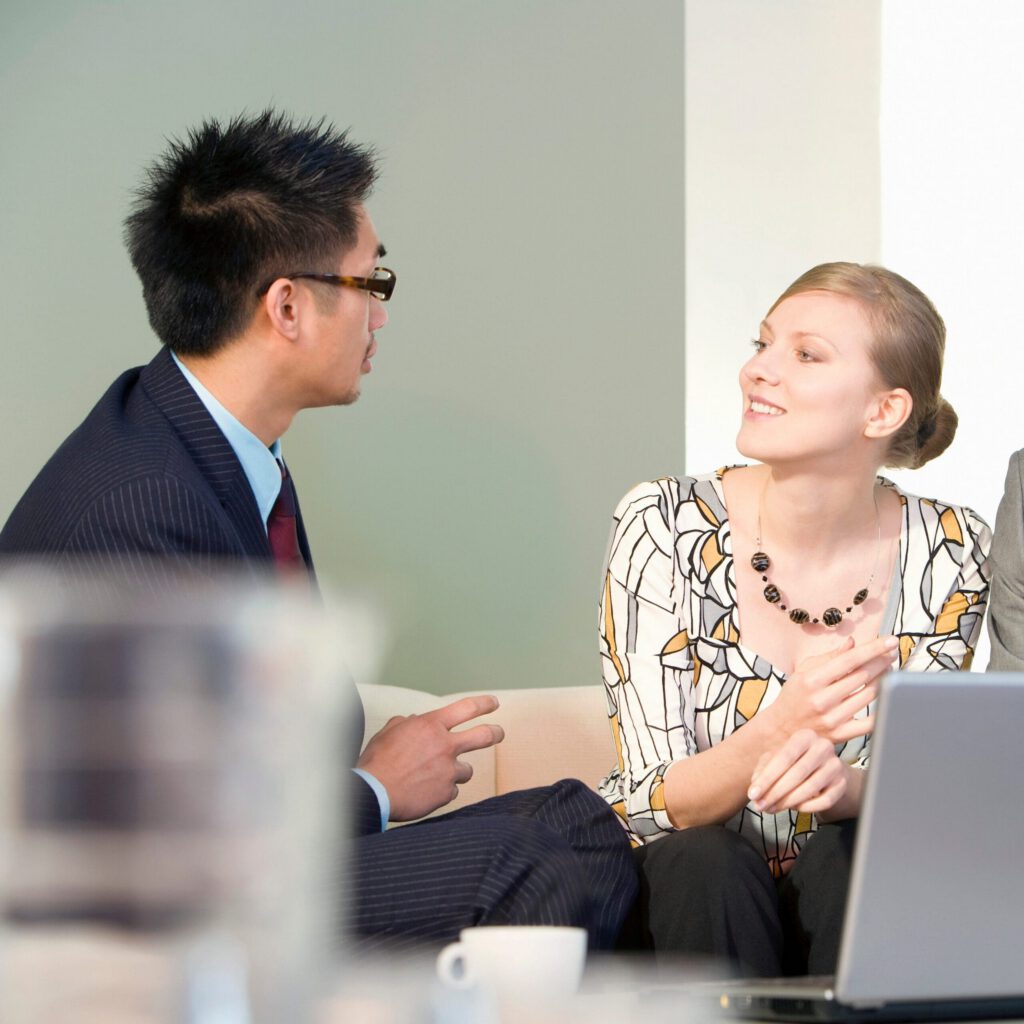 Two people in business attire sit on a couch, engaged in conversation. A laptop is open in front of them, and a cup is visible in the foreground. The woman is smiling and facing the man.