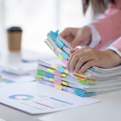 A person in a pink blazer organizes stacks of paper with colorful paper clips on a desk, with charts, documents, and a coffee cup visible in the background.