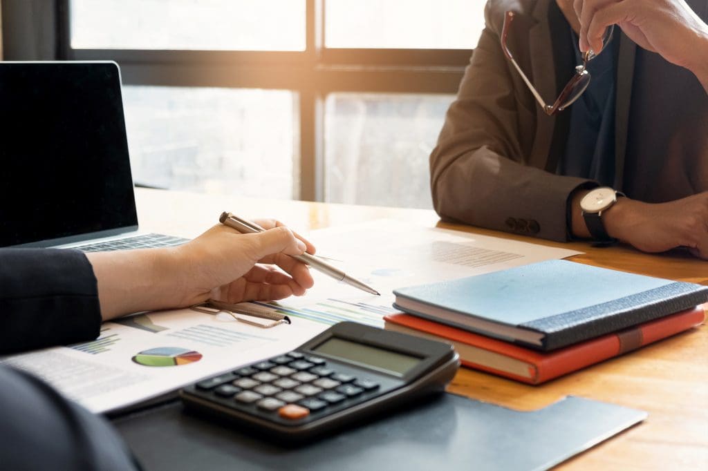 Two people sit at a desk reviewing charts and graphs, with a calculator, notebooks, laptop, and documents spread out in front of them. One person holds a pen, and the other holds glasses.