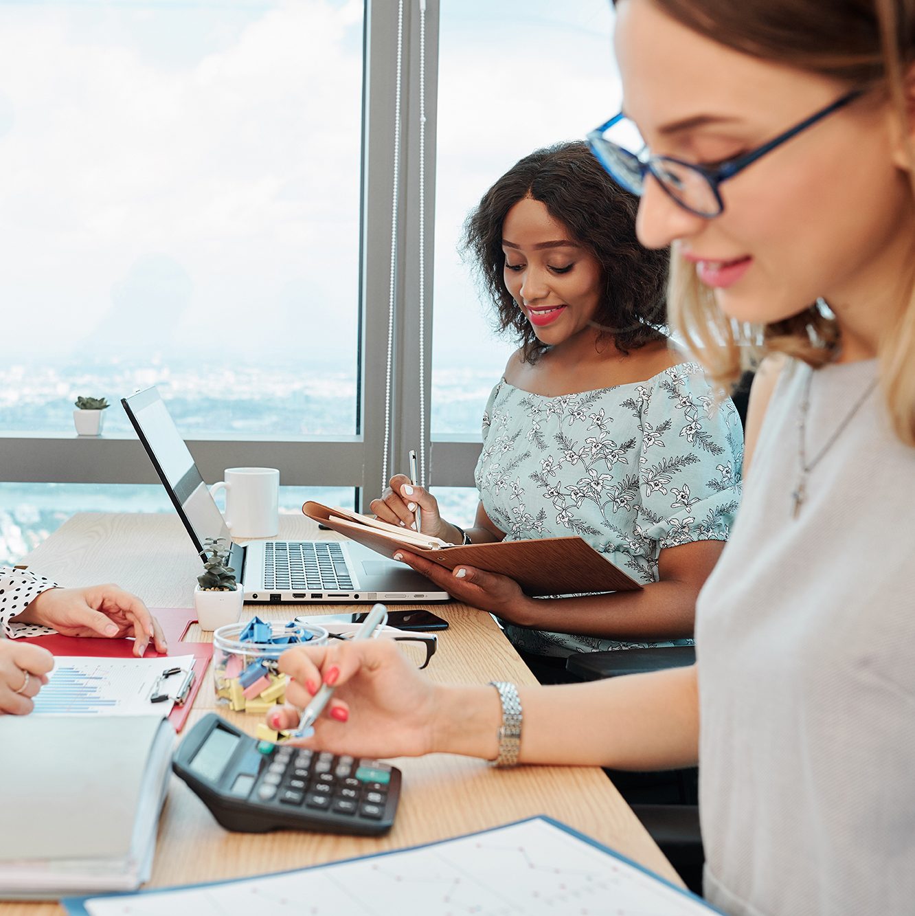 Three women work at a desk by a window with laptops, papers, and a calculator; one smiles while writing on a clipboard, another types, and the third calculates.