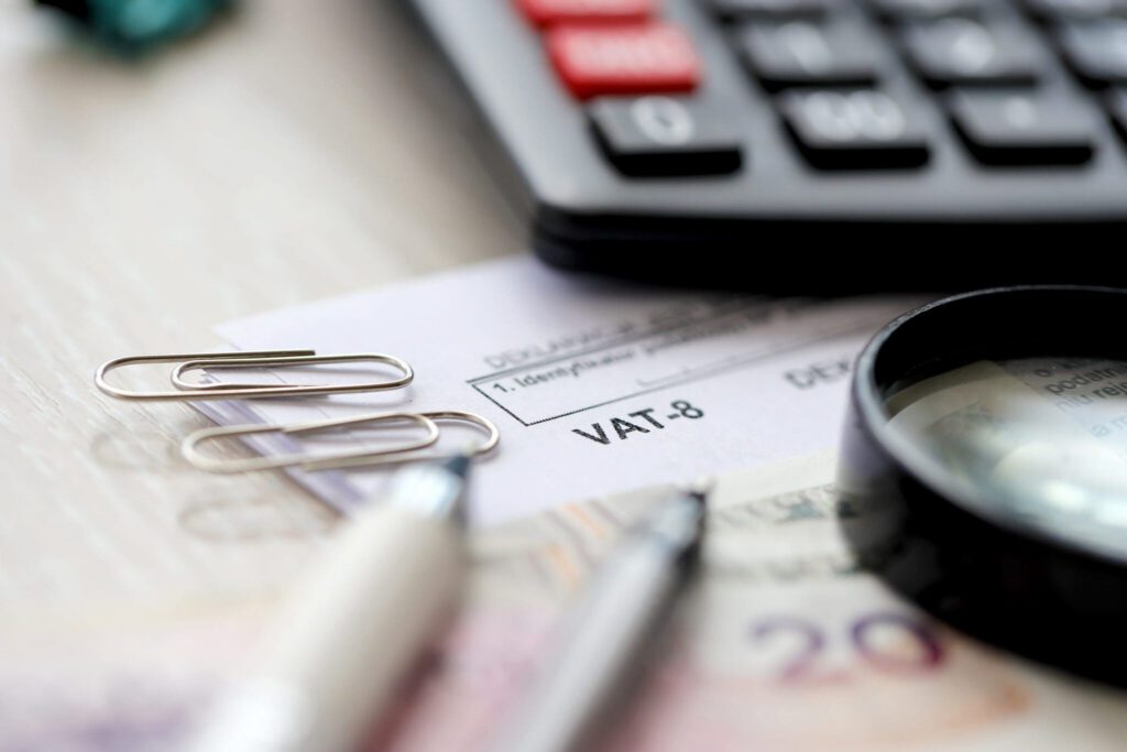 Close-up of a VAT-8 tax document on a desk with a magnifying glass, two pens, paper clips, a calculator, and part of a 20 banknote.