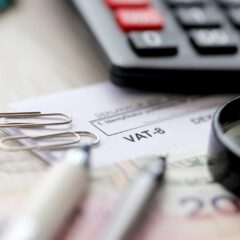 Close-up of a VAT-8 tax document on a desk with a magnifying glass, two pens, paper clips, a calculator, and part of a 20 banknote.