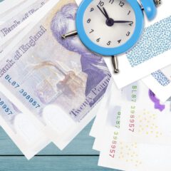 British twenty-pound notes, a blue alarm clock, a pen, and patterned envelopes are arranged on a light blue wooden surface.