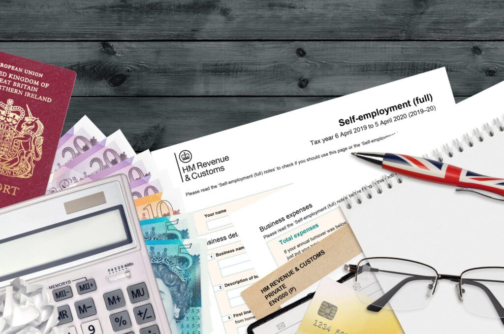 A UK passport, currency notes, a calculator, a tax form, paperclips, a pen with a Union Jack design, business receipts, and a notepad are arranged on a wooden surface.