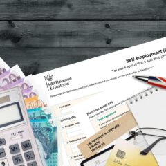 A UK passport, currency notes, a calculator, a tax form, paperclips, a pen with a Union Jack design, business receipts, and a notepad are arranged on a wooden surface.
