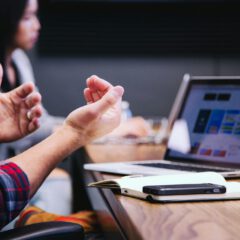 Two people sit at a table with a laptop, notebook, and phone. One person gestures with their hands while discussing something on the laptop screen. The focus is on collaboration and teamwork.
