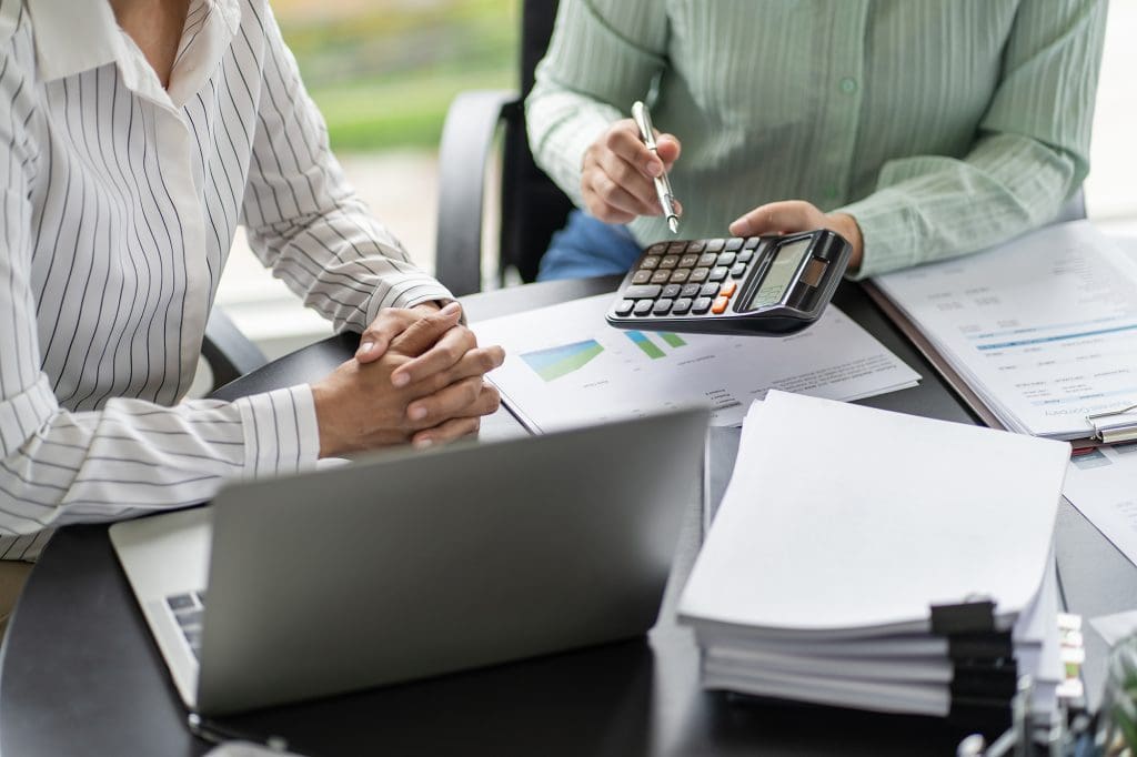 Two people sit at a desk with a laptop, papers, and charts. One points at a calculator, discussing financial data. The scene suggests a business or finance meeting.