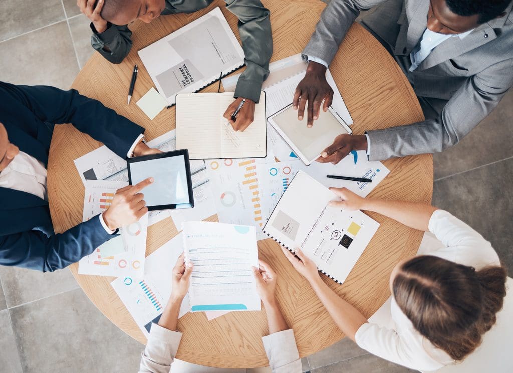 Four people sit around a round table covered with papers, charts, a tablet, notebooks, and a laptop, collaborating and discussing work.