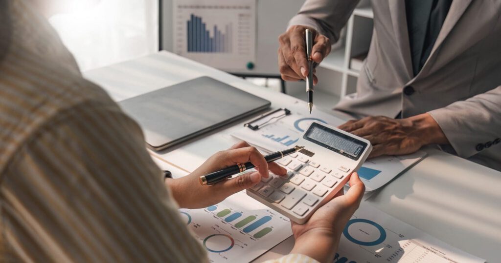 Two people discuss financial charts at a desk, one holding a calculator and a pen, the other pointing at a document with graphs. A laptop and more charts are visible in the background.