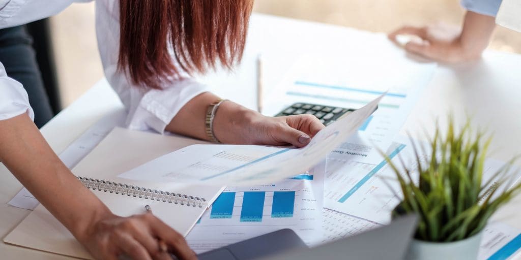 Two people review financial charts and documents on a white desk with a notebook, calculator, and a small green plant in the foreground.